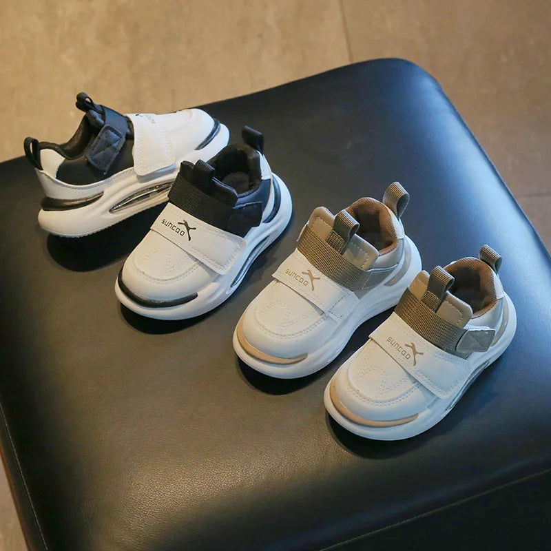 A pair of white and black kids' everyday sneakers with easy velcro straps resting on a dark surface.