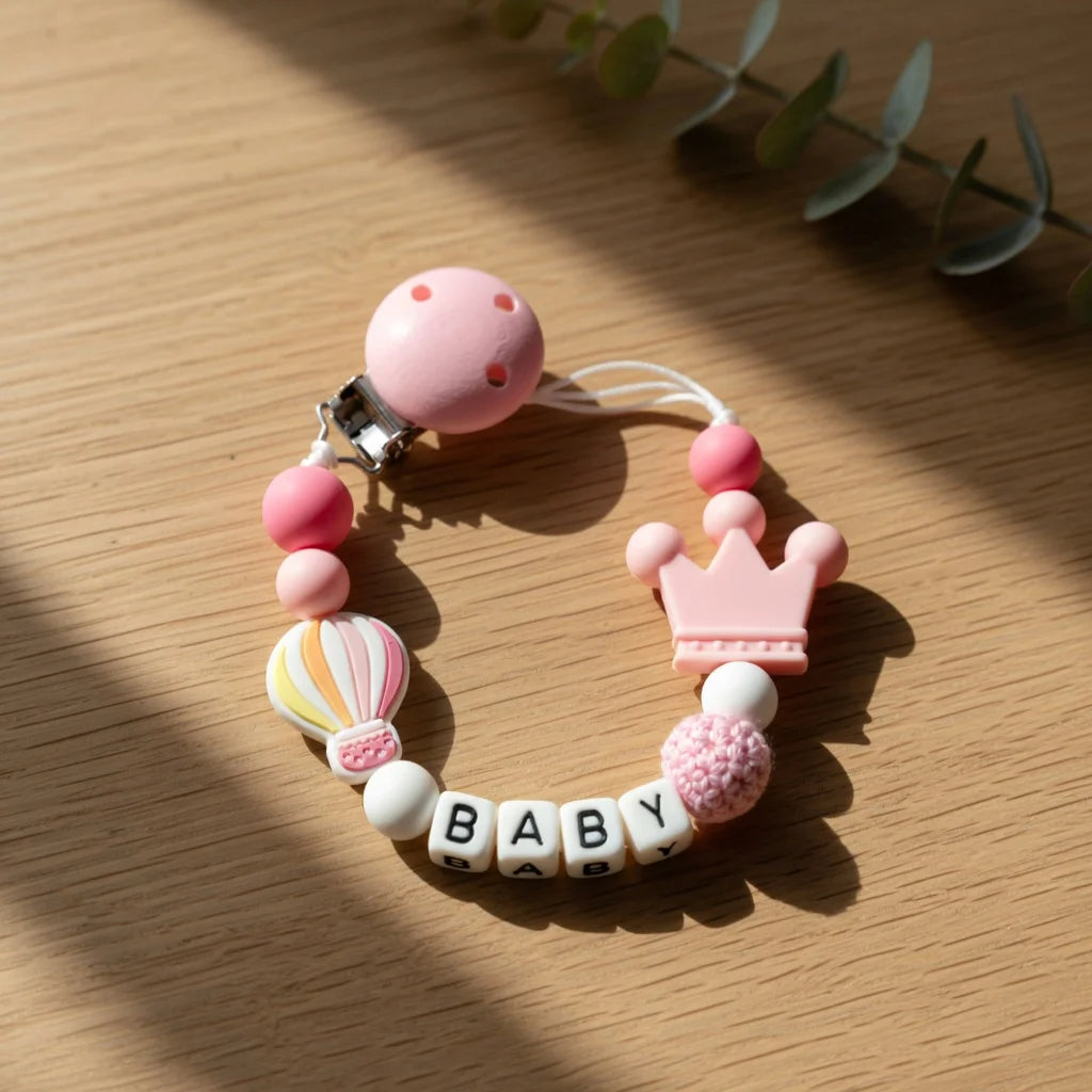A silicone pacifier clip bracelet featuring pink and white beads and a crown charm on a wooden surface.