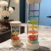 A clear wooden rain sound shaker toy with colorful plastic elements stacked inside a tall wooden holder.