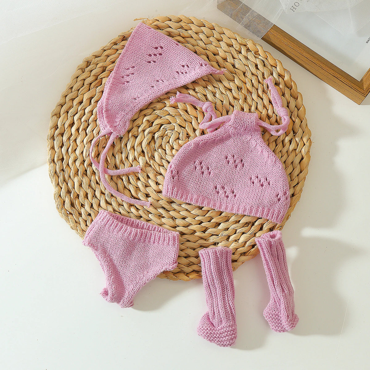 A pink knitted doll outfit set for a baby doll featuring a hat, top, and leg warmers, displayed on a woven mat.