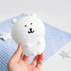 A hand holds a white silicone lamp shaped like a bear with black facial features on a blue checkered cloth.