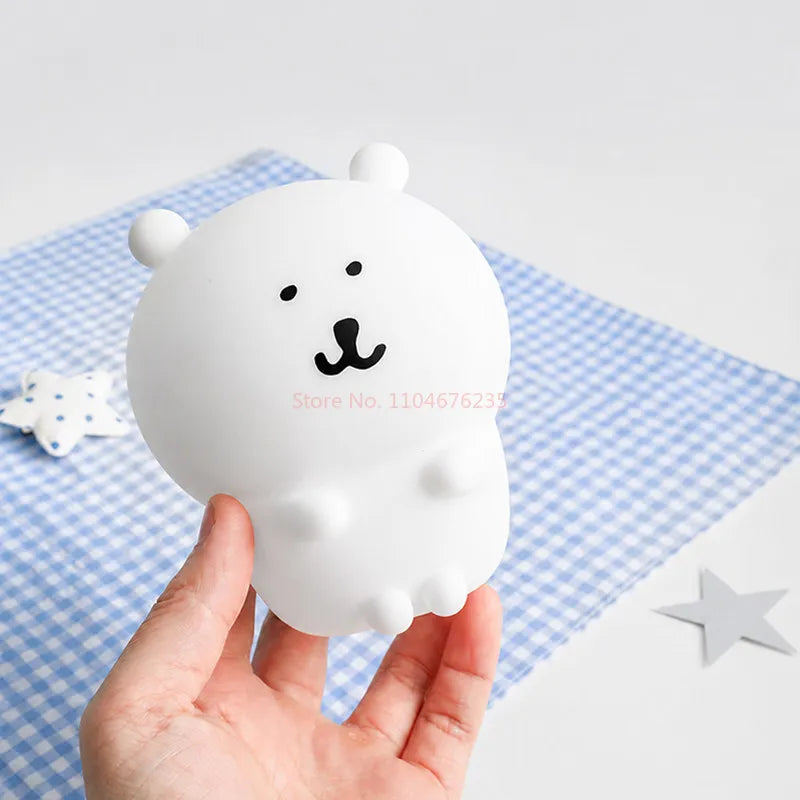 A hand holds a white silicone lamp shaped like a bear with black facial features on a blue checkered cloth.