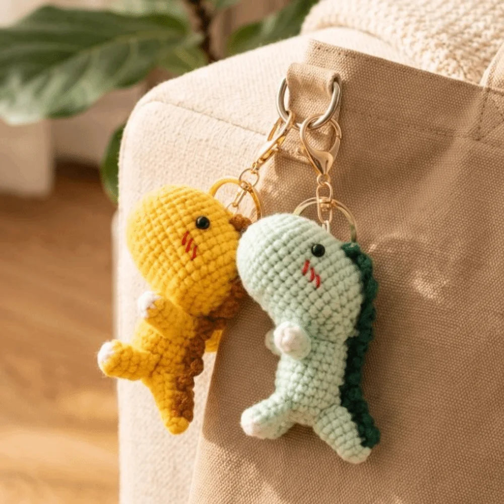A handmade crochet yellow dinosaur and a light green dinosaur keychain hanging from a gold clasp on a beige canvas bag.