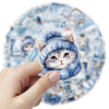 A hand holds a blue knitted cat sticker featuring white fur and large ears against a light blue watercolor background.