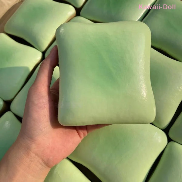 A hand holds a square, pale green sensory stress relief clay toy.