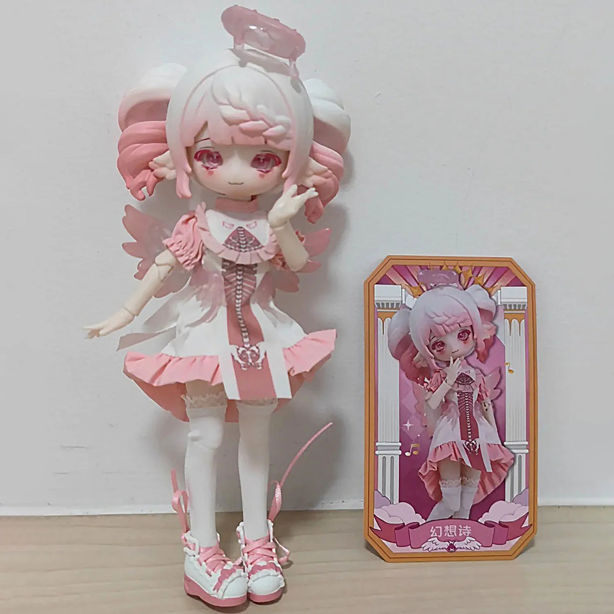 A pink and white articulated BJD doll with pink hair and wings standing next to its packaging box.