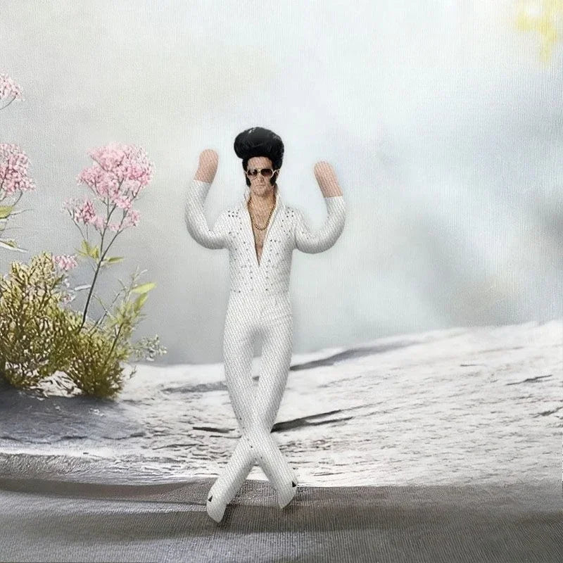 A white plush doll dressed as Elvis Presley stands on a gray surface with pink flowers in the background.