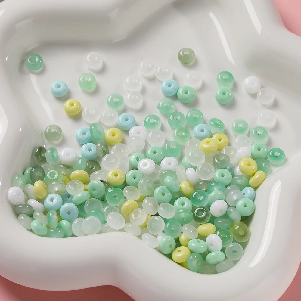 A collection of small, round imitation jade glass spacer beads in various shades of green and white are displayed in a white 