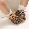 A pair of leopard print soft sole baby sneakers worn by a doll's foot.