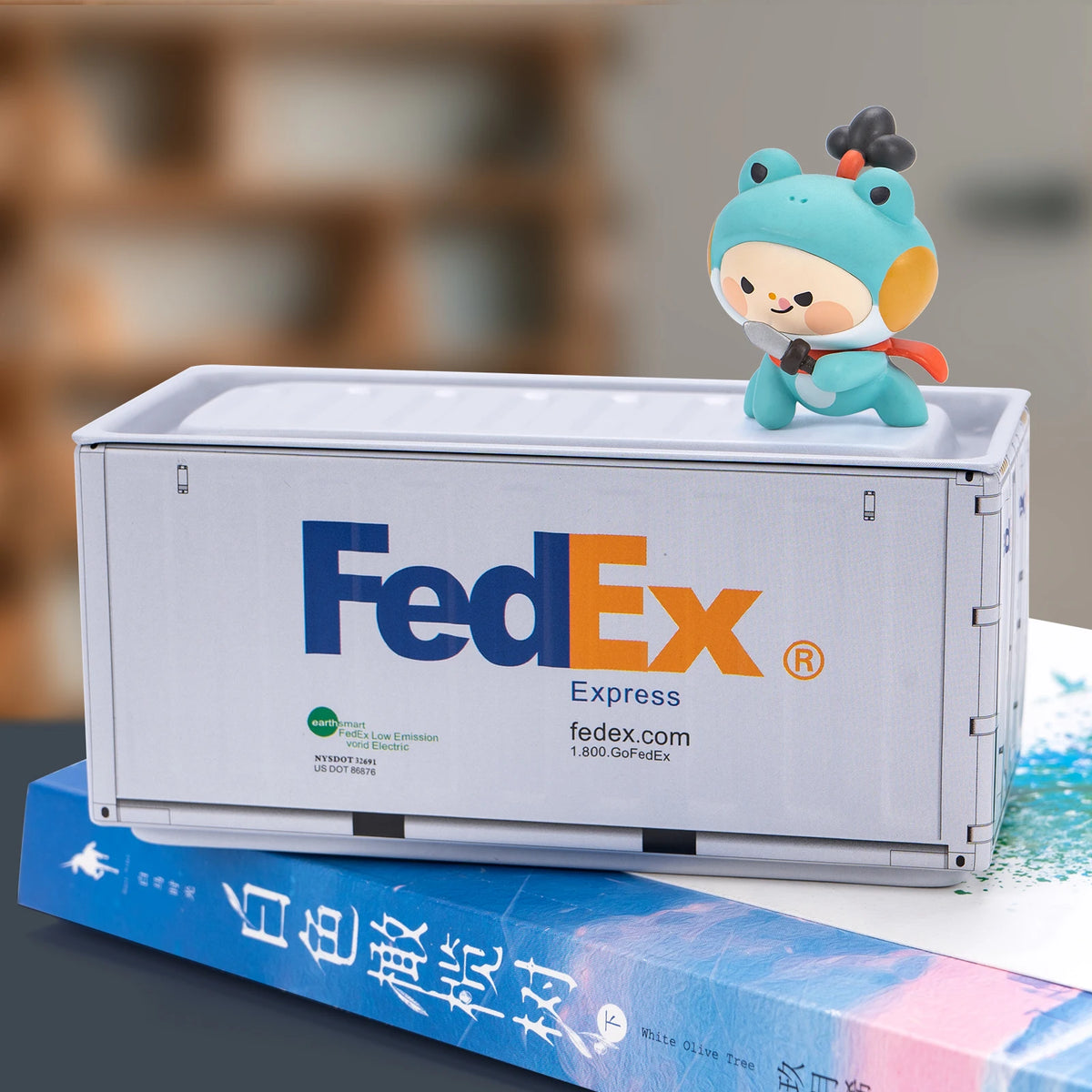 A small, light blue figurine sits on top of a white FedEx industrial storage box.