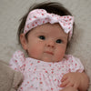 A close-up photo of a baby doll with hand-rooted hair wearing a pink floral outfit and a matching bow.