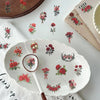 A white plate with a floral pattern and a wooden spoon rests on a white cloth, surrounded by vintage red rose stickers.