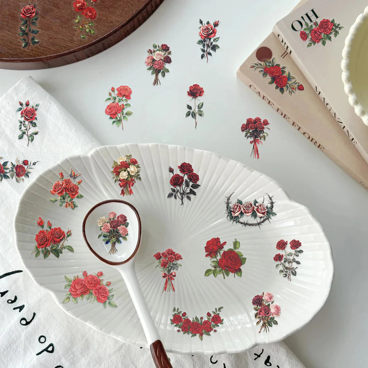 A white plate with a floral pattern and a wooden spoon rests on a white cloth, surrounded by vintage red rose stickers.