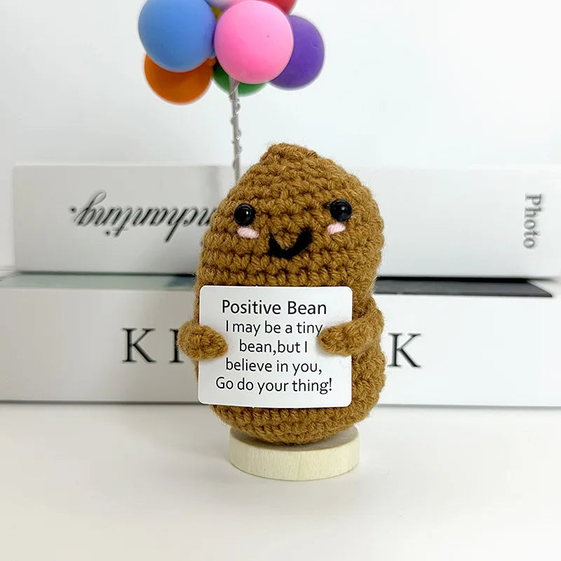 A handwoven crochet ornament shaped like a bean with a smiling face, sitting on a wooden base.