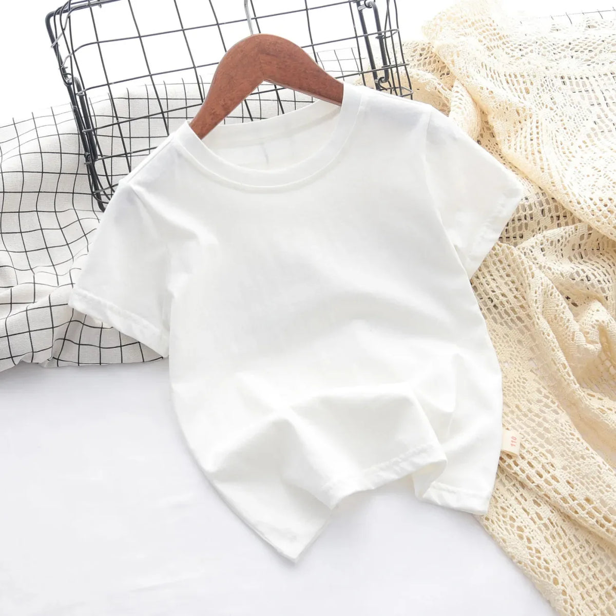 A white classic cotton crew neck t-shirt for kids is displayed on a white surface next to a mesh basket and a beige knitted f