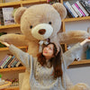 A young woman is hugging a giant, light brown plush teddy bear in a cozy indoor setting.