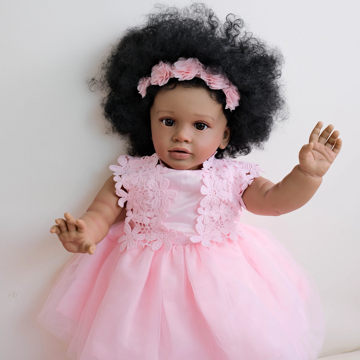 A lifelike reborn toddler doll with hand-rooted black curly hair wearing a pink lace dress.