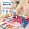 A hand is painting a diamond art canvas with a pen, surrounded by various colored beads and paint palettes.
