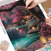 A close-up of a diamond painting kit featuring a vibrant pink and green landscape with a flowing river and traditional archit