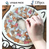 A hand is placing a piece of a cat-shaped wooden jigsaw puzzle onto the board, showing 135 unique pieces.