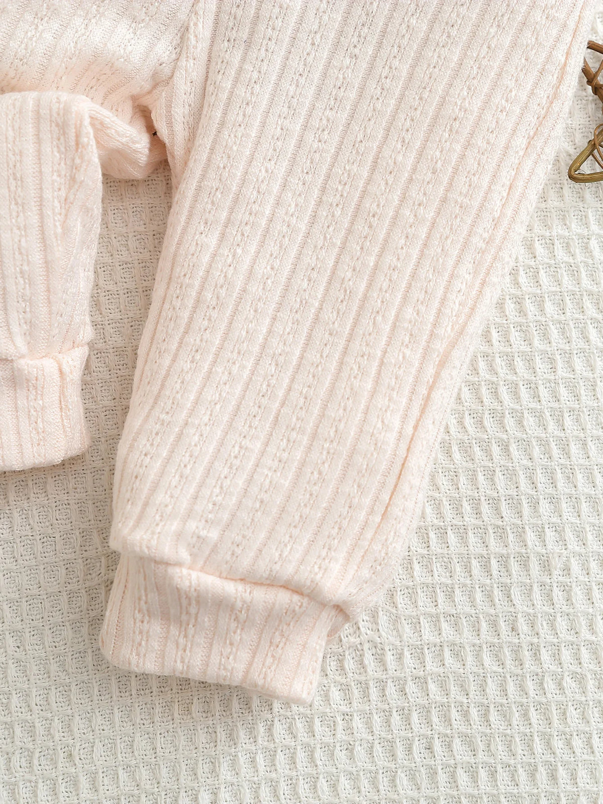A light pink knitted baby romper and pants set laid flat on a white textured fabric.