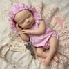 A newborn silicone reborn baby doll with pink lace bonnet lying on a pink crocheted fabric.