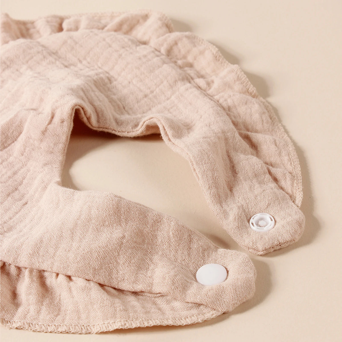 A light pink cotton ruffle baby bib is shown flat on a beige background.
