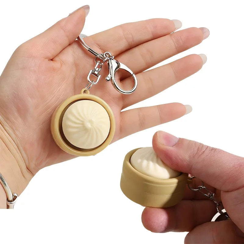A hand holds a keychain featuring a small, round, beige steamed bun-shaped fidget toy.