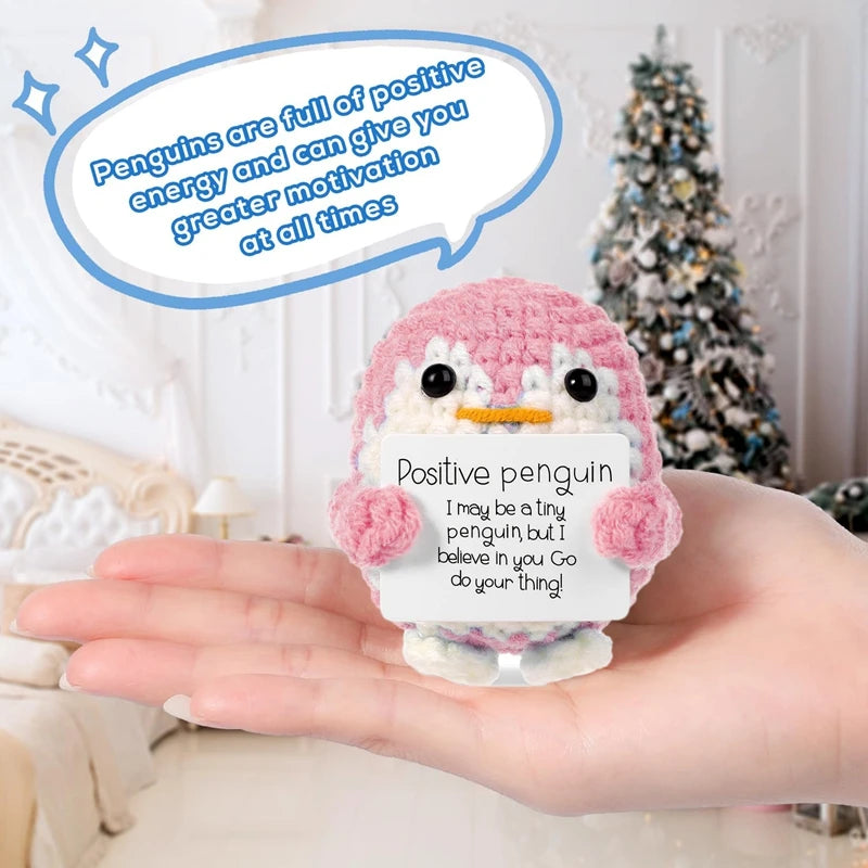 A hand holds a handmade pink crochet penguin doll with a positive message card, set against a blurred background.