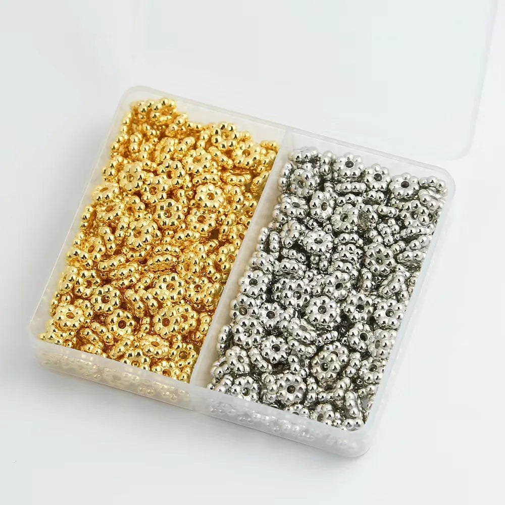 A clear plastic box containing two piles of small silver and gold snowflake spacer beads for jewelry making