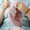 Hands holding a pink knitted item with colorful locking stitch markers on a light blue background.
