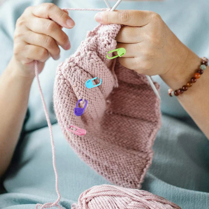Hands holding a pink knitted item with colorful locking stitch markers on a light blue background.