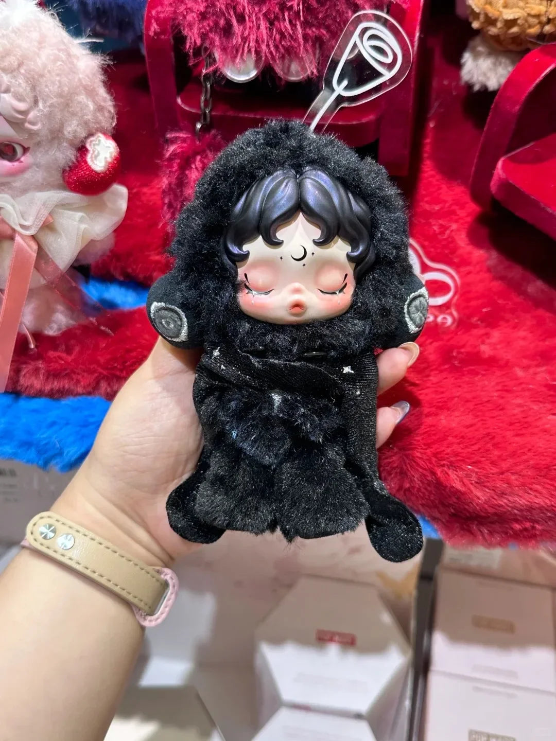 A hand holds a black Skullpanda You Found Me! Series collectible figure with dark hair and closed eyes against a red fuzzy ba