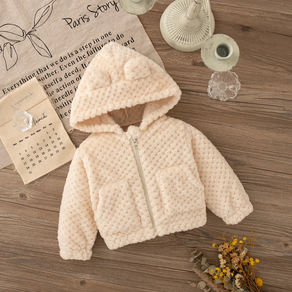 A light pink plush hooded winter jacket lying on a wooden surface next to a notebook and a woven object.