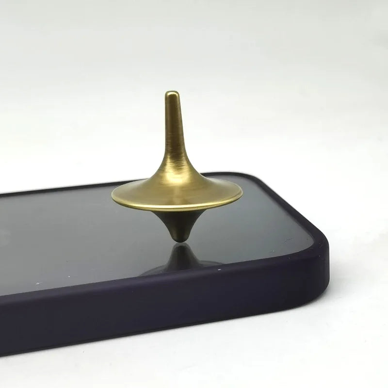A brass metal fidget spinner with a smooth, tapered top resting on a dark rectangular surface.