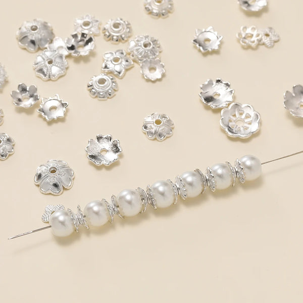 Silver alloy flower shape bead caps scattered on a light background with a string of pearl beads below