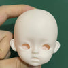 A light pink 1/6 scale BJD doll head with sculpted features being held by a hand against a dark green background.