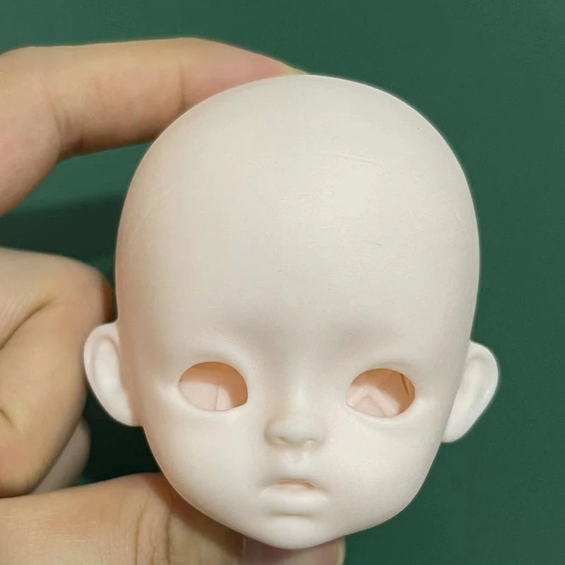 A light pink 1/6 scale BJD doll head with sculpted features being held by a hand against a dark green background.