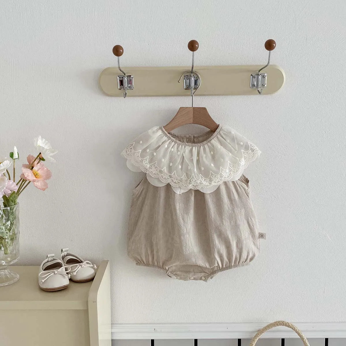 A beige sleeveless cotton jumpsuit for a baby girl with a white lace collar hanging on a wall rack.