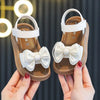 A pair of beige sandals with white bow details being held by a person against a dark background.