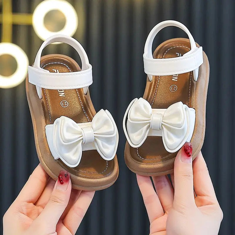 A pair of beige sandals with white bow details being held by a person against a dark background.