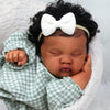 A newborn baby doll with hand-rooted hair wearing a white bow and a green checkered outfit.