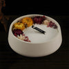 White candle in a round ceramic dish surrounded by dried flowers and leaves on a dark wooden surface