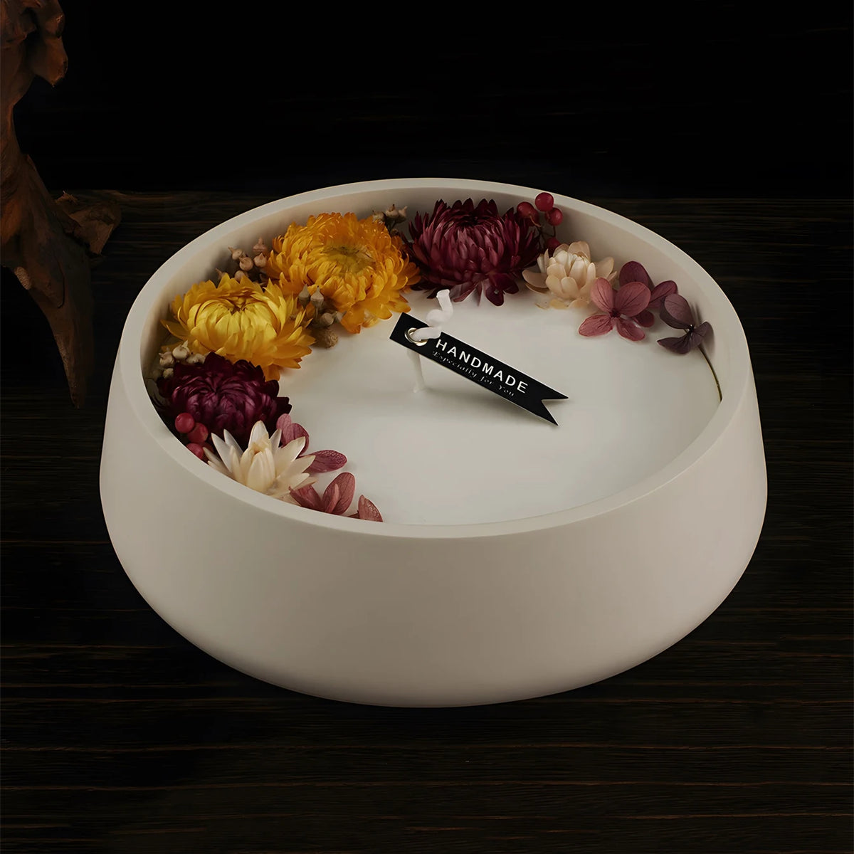 White candle in a round ceramic dish surrounded by dried flowers and leaves on a dark wooden surface