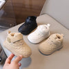 A hand holds a pair of beige quilted toddler winter snow boots with soft soles and Velcro closures.