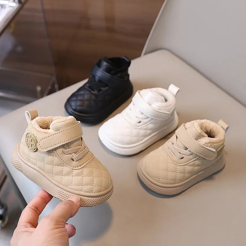A hand holds a pair of beige quilted toddler winter snow boots with soft soles and Velcro closures.