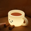 A cute, glowing coffee cup shaped night light with a smiling face rests on a brown surface.