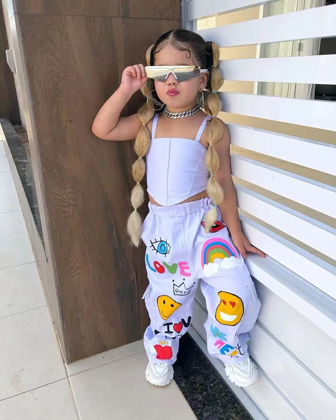 A young girl wearing a white two-piece outfit with suspender top and long pants, accessorized with sunglasses and jewelry.