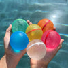 A hand holds several colorful reusable water balloons including green, yellow, orange, blue, and pink spheres in clear water.