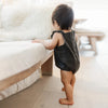A toddler wearing a dark denim romper stands next to a light-colored sofa.
